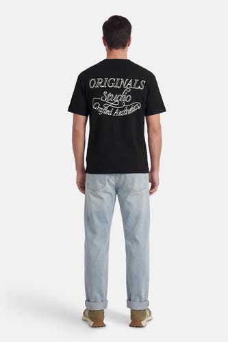 T-shirt noir à manches courtes de ORIGINALS BY JACK & JONES, avec imprimé texte blanc au dos, combiné avec un jean bleu clair et des baskets vertes.