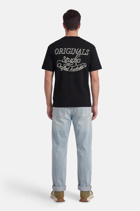 T-shirt noir à manches courtes de ORIGINALS BY JACK & JONES, avec imprimé texte blanc au dos, combiné avec un jean bleu clair et des baskets vertes.