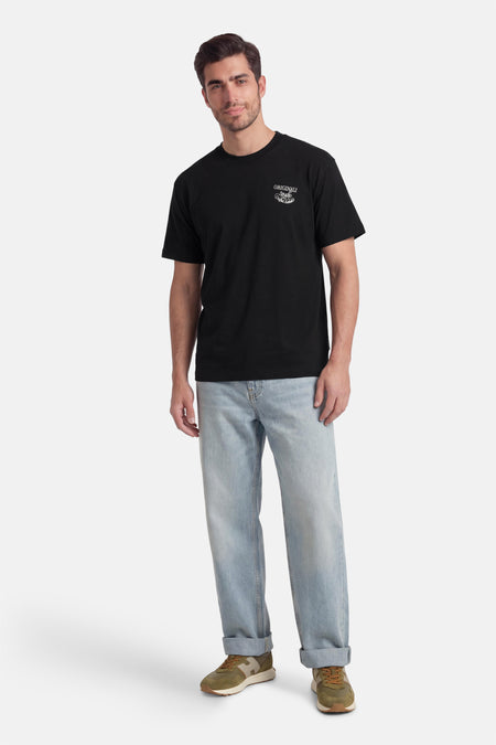 T-shirt noir à manches courtes de ORIGINALS BY JACK & JONES, avec un petit imprimé blanc sur la poitrine, porté avec un jean bleu clair et des baskets vertes.