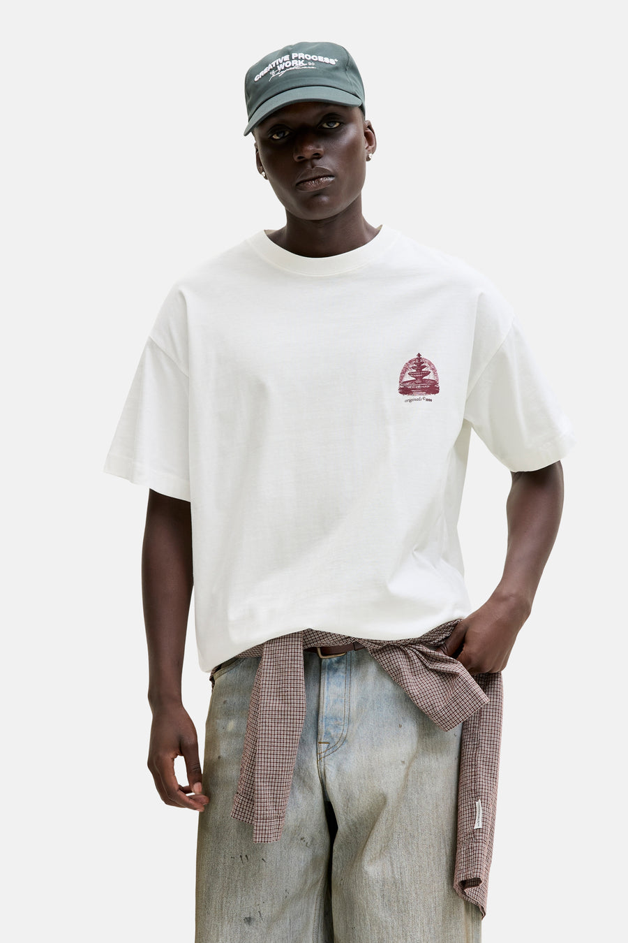 T-shirt blanc de ORIGINALS BY JACK & JONES, avec un petit imprimé rouge sur la poitrine, porté avec un jean ample et une chemise à carreaux à la taille.