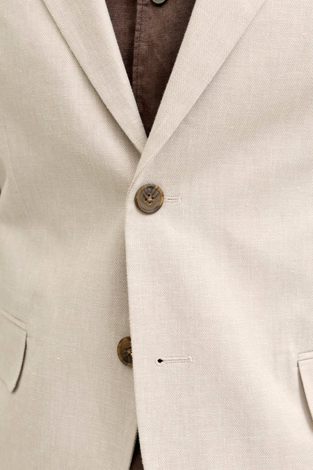 Détail d'un blazer écru de PREMIUM by JACK & JONES en tissu texturé, avec des boutons marron et une chemise marron en dessous.