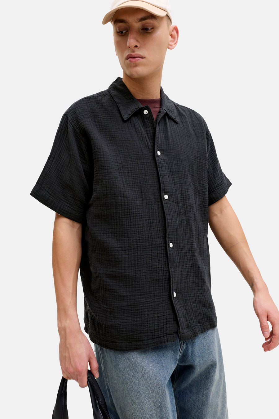 Chemise noire à manches courtes de ORIGINALS BY JACK & JONES, avec texture et fermeture à boutons, portée avec un jean et une casquette beige.