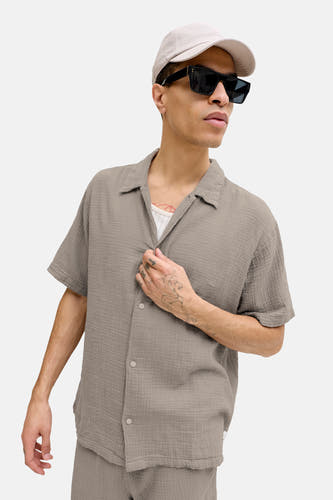Chemise marron à manches courtes de ORIGINALS BY JACK & JONES, avec boutons, portée avec une casquette et des lunettes de soleil.