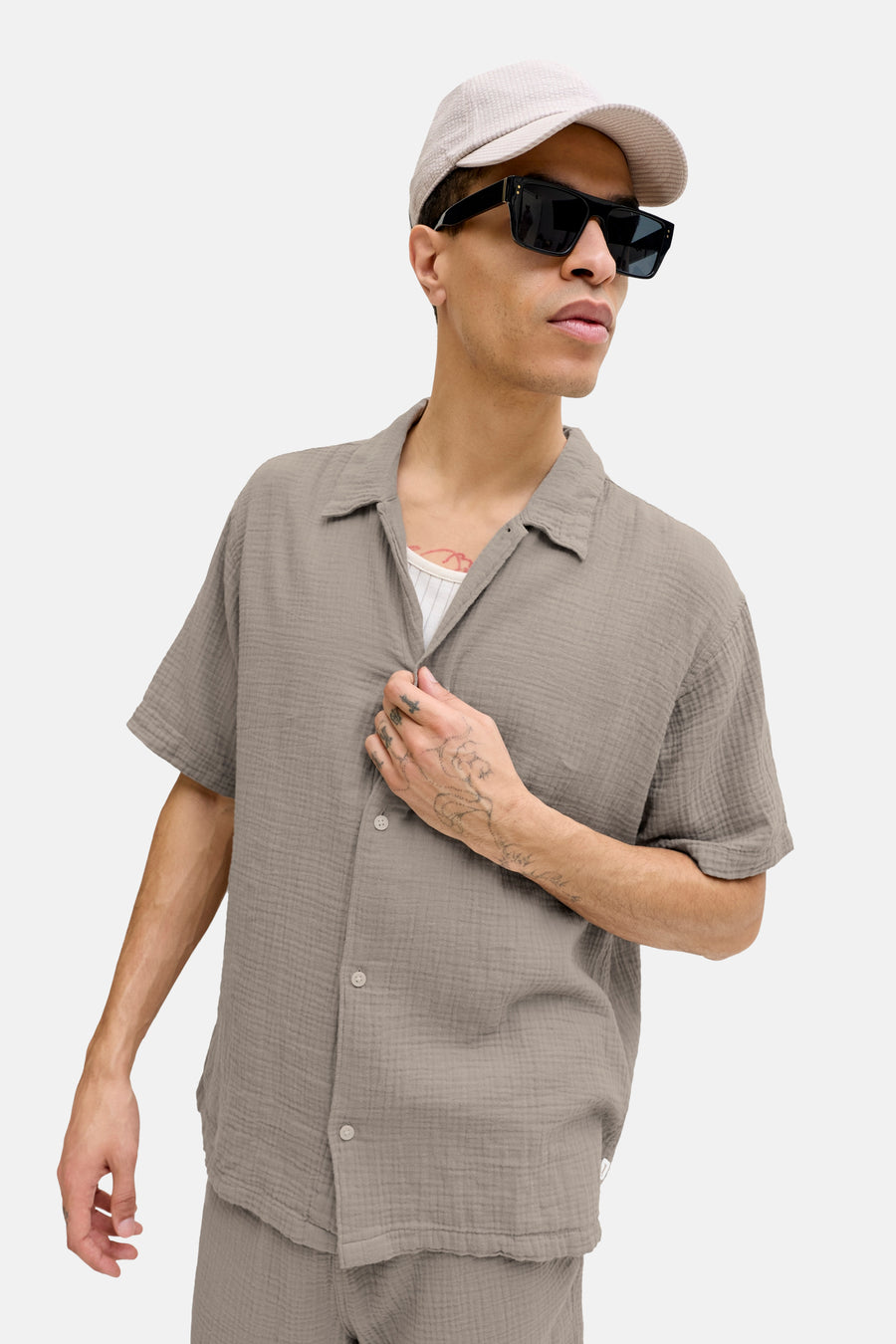 Chemise marron à manches courtes de ORIGINALS BY JACK & JONES, avec boutons, portée avec une casquette et des lunettes de soleil.