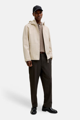 Korte ecru jas van PREMIUM by JACK & JONES, met capuchon, gedragen over een bruine polo en donkere pantalon.
