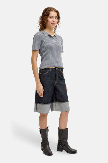 Donkerblauwe denim short van JJXX, met omgeslagen zoom, gecombineerd met een grijs gestreepte polo en bruine laarzen.