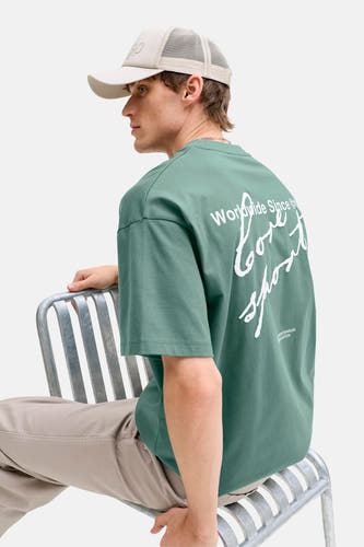 T-shirt vert de CORE BY JACK & JONES avec imprimé blanc dans le dos, porté avec une casquette et un pantalon beiges.
