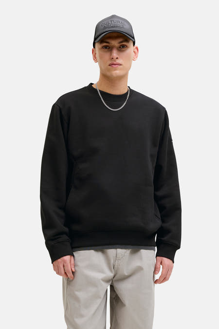 Pull noir de CORE BY JACK & JONES, avec un col rond et des manches longues, porté avec une chaîne argentée et un pantalon gris.