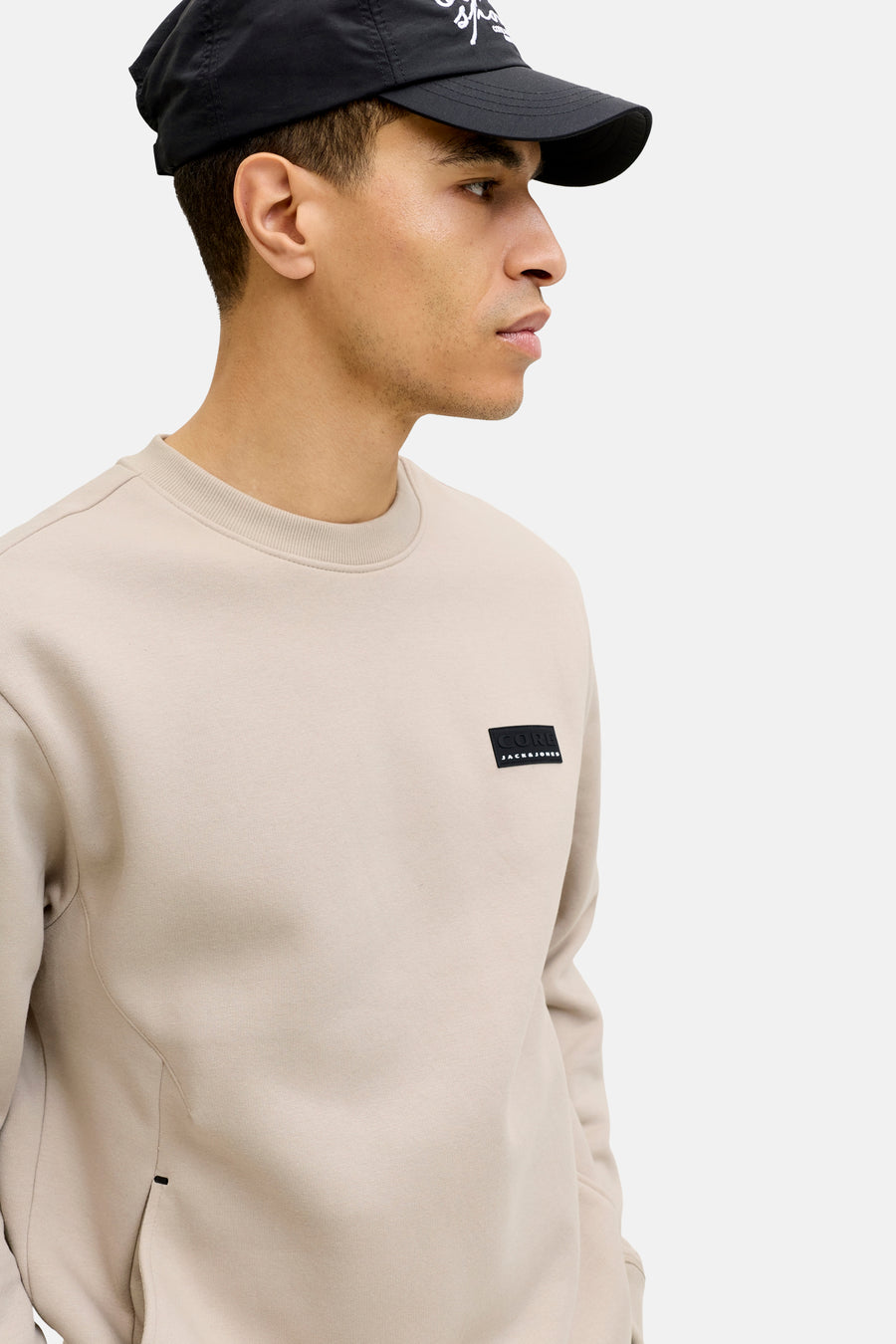 Pull beige de CORE by JACK & JONES, avec un patch logo noir sur la poitrine et une casquette noire.
