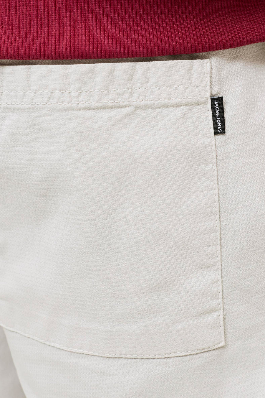 Short beige de JACK & JONES JEANS INTELLIGENCE avec une texture tissée, une poche visible avec étiquette noire et un tissu côtelé rouge au-dessus.