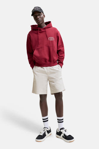 Short beige de JACK & JONES JEANS INTELLIGENCE, porté par un mannequin avec un sweat à capuche rouge, une casquette noire et des baskets.