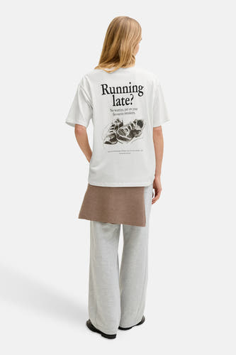 T-shirt blanc à manches courtes de JJXX, avec texte noir et illustration de chaussure au dos, porté avec un pantalon gris.