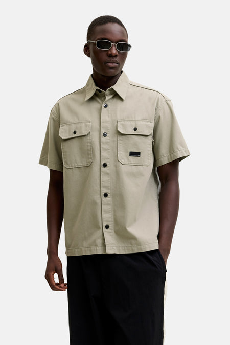 Chemise beige à manches courtes de CORE BY JACK & JONES, avec poches poitrine et étiquette noire, portée par un homme avec des lunettes de soleil et un pantalon noir.