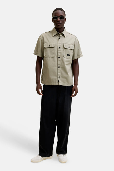 Chemise beige à manches courtes de CORE BY JACK & JONES, avec deux poches poitrine, boutons noirs et un écusson, portée avec un pantalon noir et des baskets blanches.