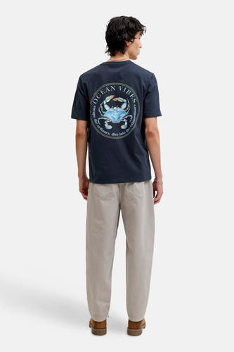 T-shirt bleu à manches courtes de ORIGINALS BY JACK & JONES, avec un imprimé rond "Ocean Vibes" dans le dos, porté avec un pantalon beige et des chaussures marron.