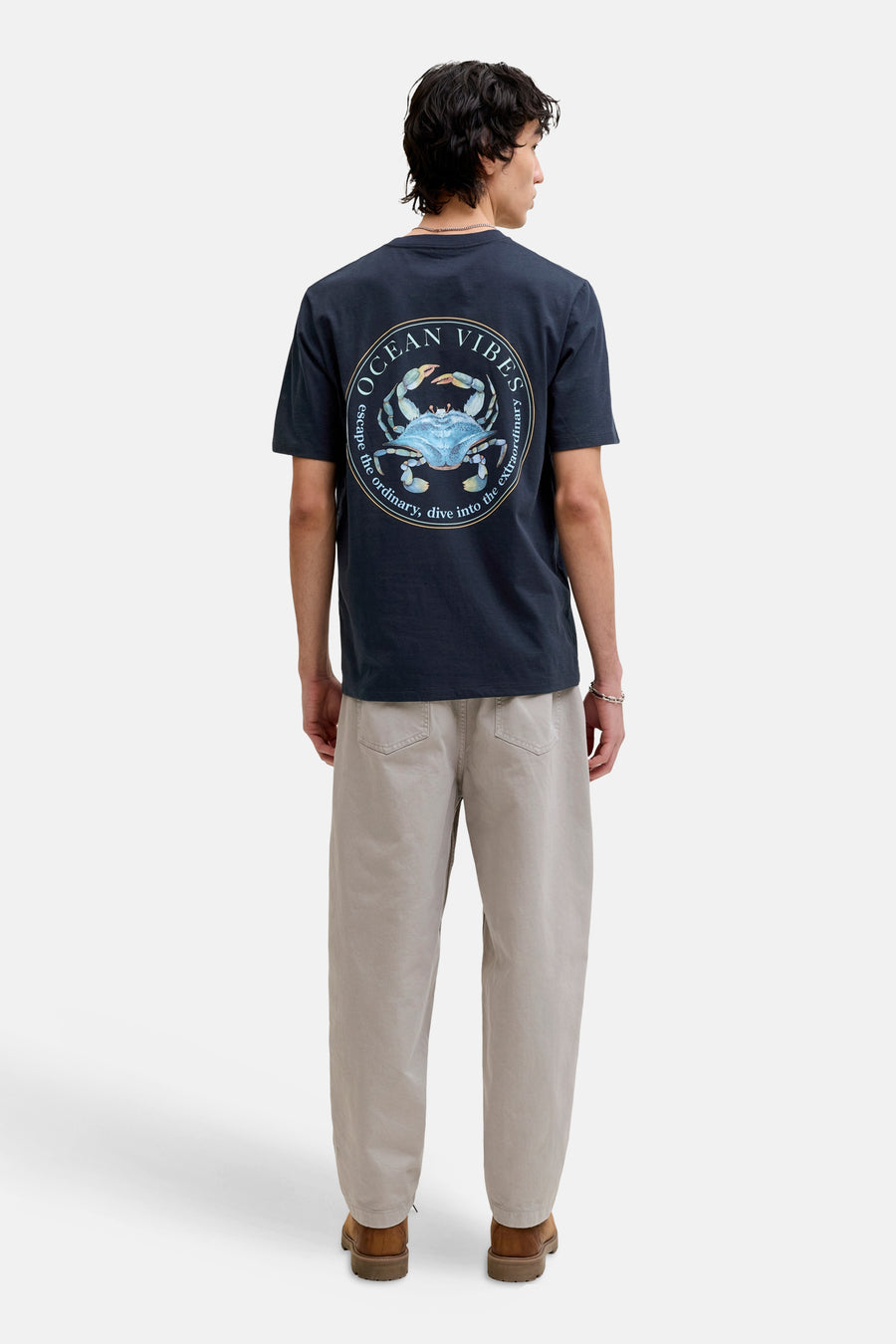 T-shirt bleu à manches courtes de ORIGINALS BY JACK & JONES, avec un imprimé rond "Ocean Vibes" dans le dos, porté avec un pantalon beige et des chaussures marron.