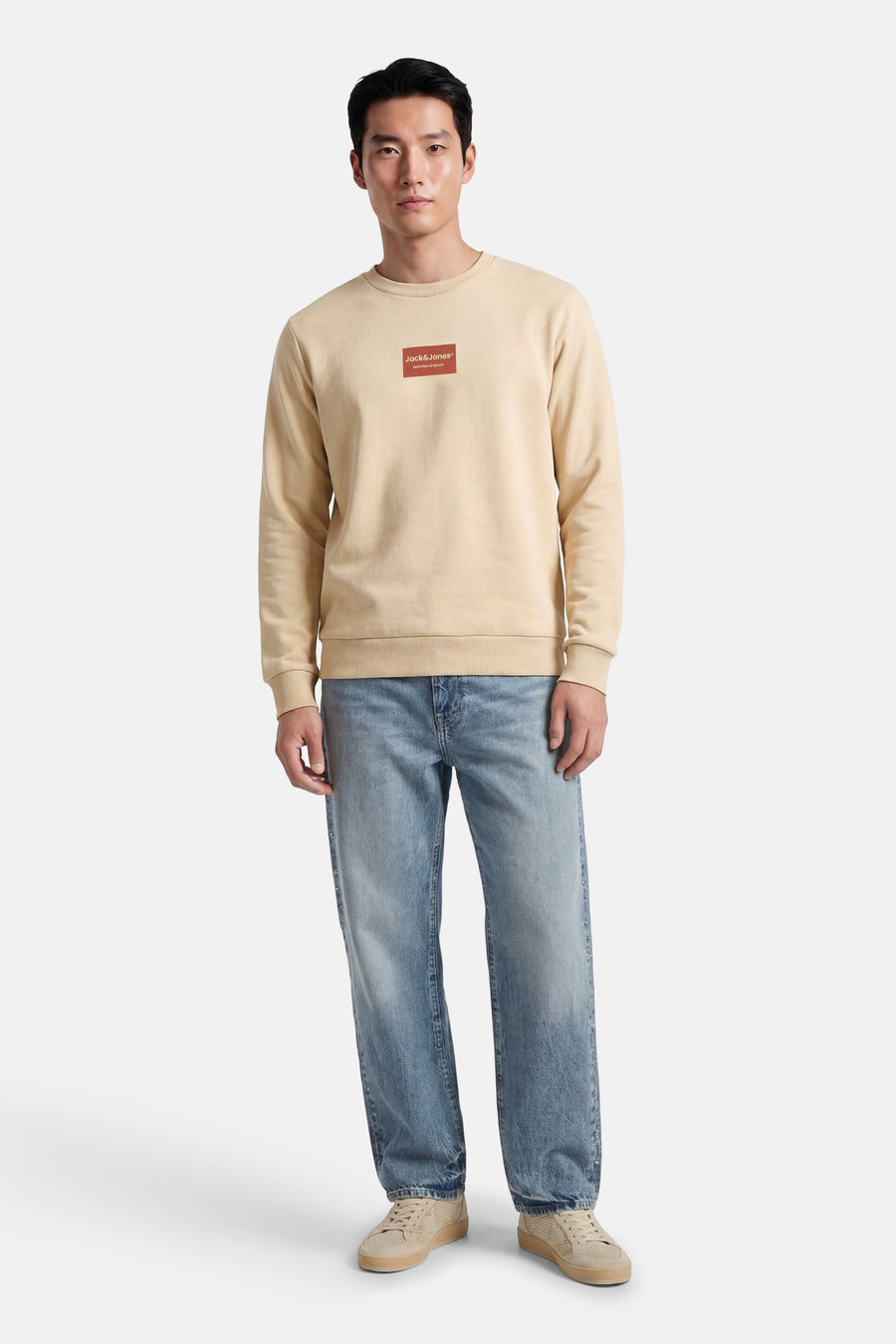 Pull beige à col rond de CORE by JACK & JONES, avec un logo rouge sur la poitrine, combiné avec un jean bleu et des chaussures beiges.
