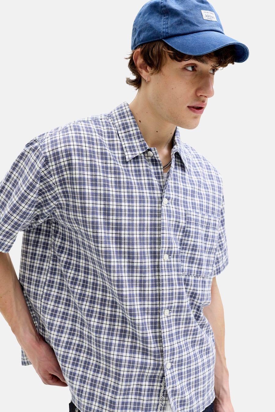 Chemise à carreaux bleus à manches courtes de ORIGINALS BY JACK & JONES, avec boutons blancs, portée par un mannequin avec une casquette bleue et une boucle d'oreille argentée.