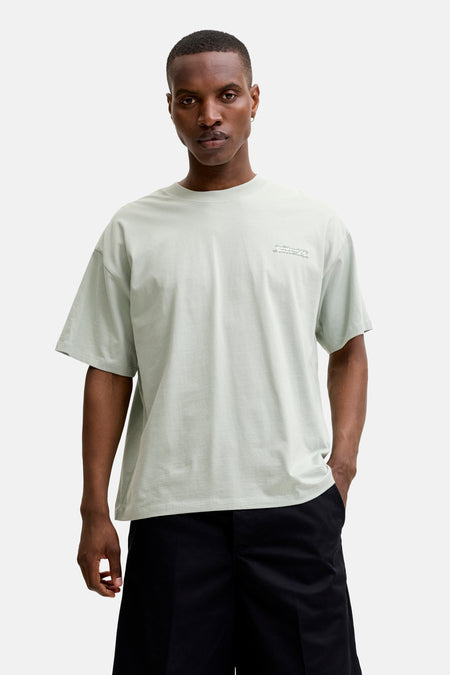 T-shirt à manches courtes vert de CORE BY JACK & JONES avec imprimé graphique gris clair sur la poitrine, stylisé avec un short noir, des chaussettes blanches à rayures noires et des baskets.