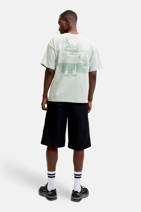 T-shirt à manches courtes vert de CORE BY JACK & JONES, avec imprimé 'Wellness Club' et 'CORE™ ISLAND RETREAT' dans le dos, porté par un homme avec un short noir et des baskets.