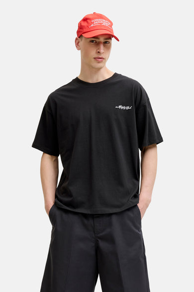 T-shirt noir à manches courtes de CORE BY JACK & JONES, avec le texte blanc 'wet 1999 Club' sur la poitrine, porté par un homme avec une casquette rouge et un short noir.