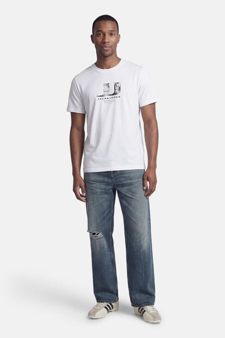 T-shirt blanc à manches courtes de CORE BY JACK & JONES, avec imprimé sur la poitrine, porté avec un jean bleu et des baskets.
