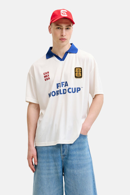 Polo blanche ORIGINALS BY JACK & JONES avec col bleu, imprimé FIFA World Cup et 'CAN MEX USA', combinée avec un short en jean et une casquette rouge.