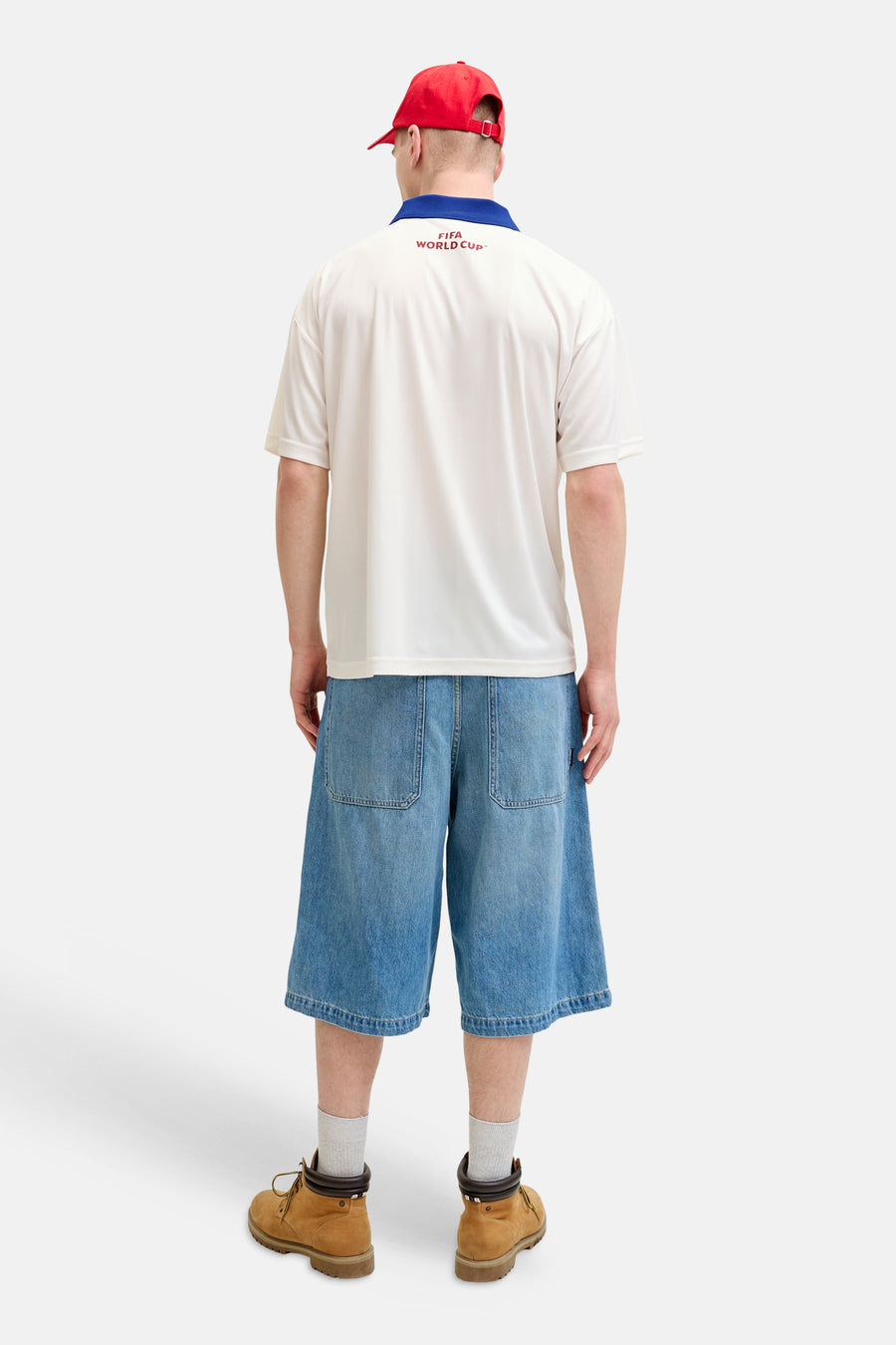 Polo blanc ORIGINALS BY JACK & JONES avec col bleu et inscription rouge 'FIFA WORLD CUP™' dans le dos, porté avec une casquette rouge et un short en jean.