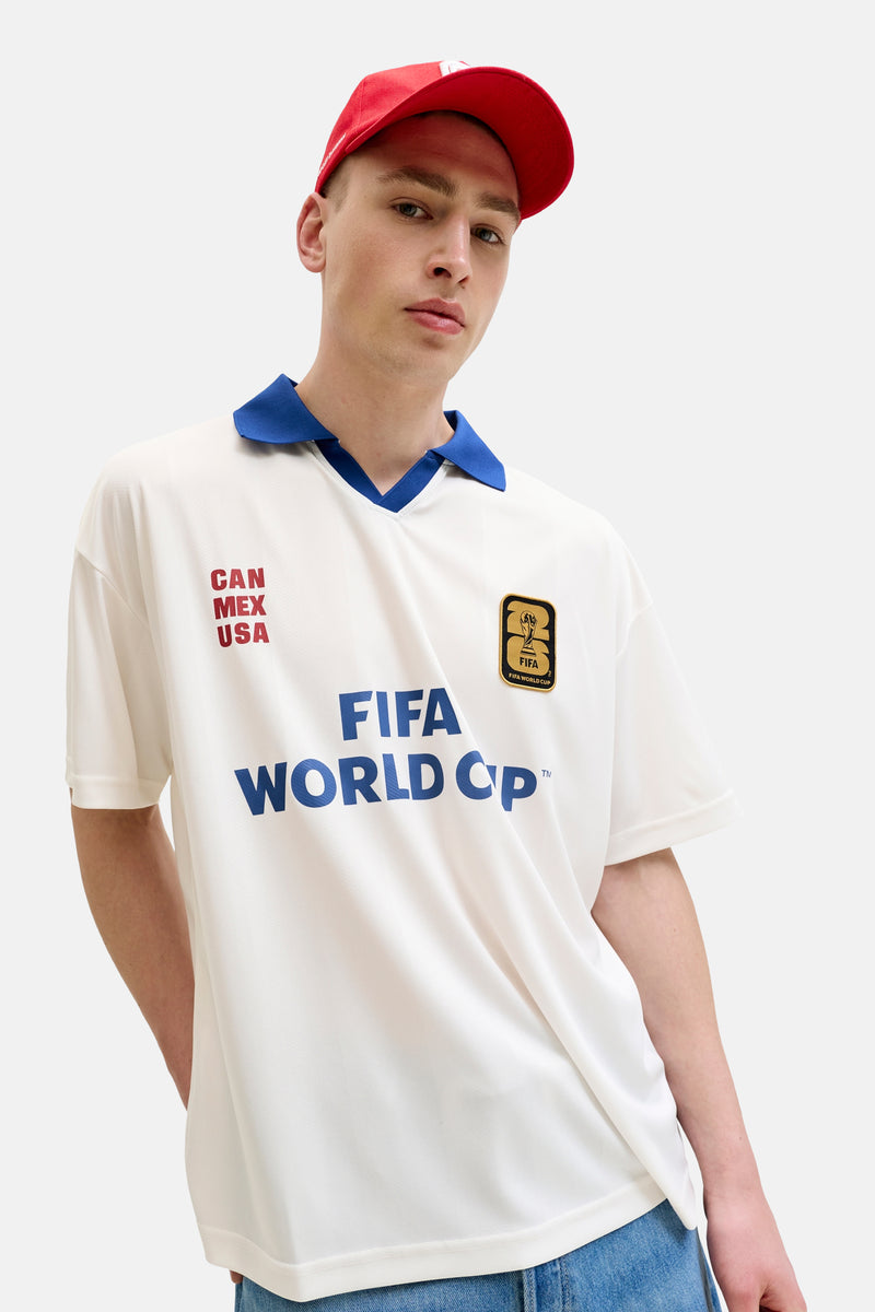 Polo blanche ORIGINALS BY JACK & JONES avec col bleu, logo FIFA World Cup 2026, textes bleus 'FIFA World Cup' et rouges 'CAN MEX USA'.