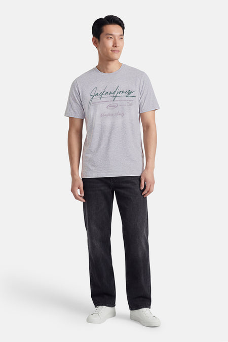 T-shirt gris de CORE BY JACK & JONES avec logo script vert, porté par un homme avec un jean noir et des baskets blanches.