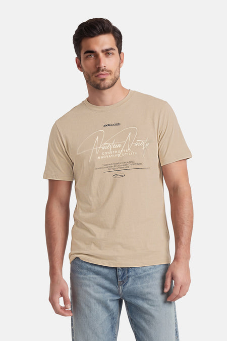 T-shirt beige de CORE BY JACK & JONES avec imprimé graphique blanc, porté par un homme en jean bleu.