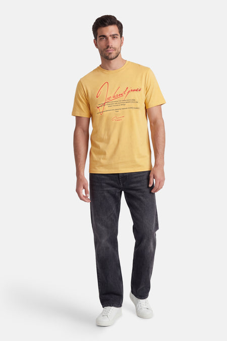 T-shirt jaune à manches courtes de CORE BY JACK & JONES avec texte rouge sur le devant, porté par un mannequin avec un jean foncé et des baskets blanches.