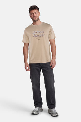 Beige T-shirt van CORE BY JACK & JONES met een gestileerde 'Jack & Jones' logo print, gedragen door een man in donkere spijkerbroek en grijze sneakers.