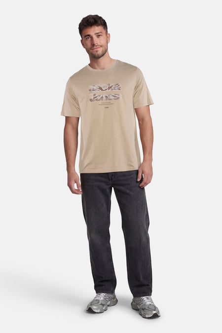 Beige T-shirt van CORE BY JACK & JONES met een gestileerde 'Jack & Jones' logo print, gedragen door een man in donkere spijkerbroek en grijze sneakers.