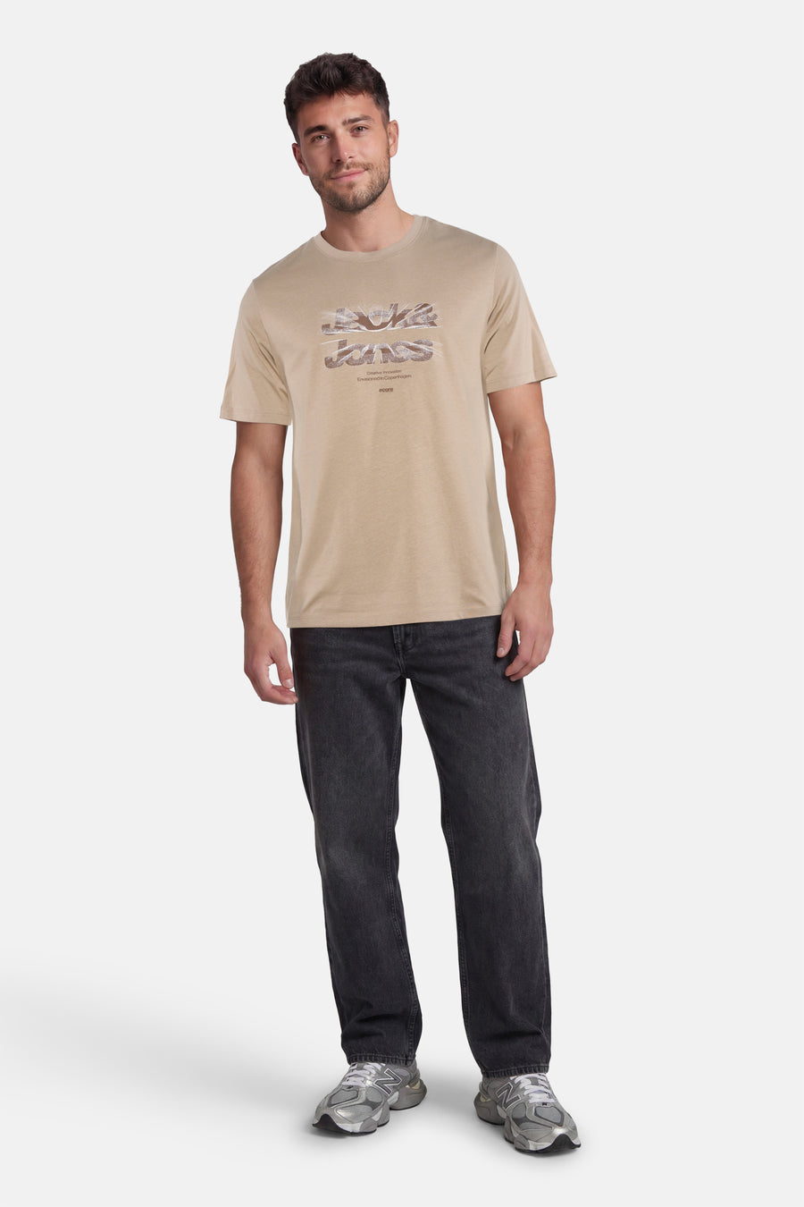 Beige T-shirt van CORE BY JACK & JONES met een gestileerde 'Jack & Jones' logo print, gedragen door een man in donkere spijkerbroek en grijze sneakers.