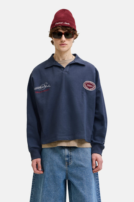 Pull bleu marine à col polo de ORIGINALS BY JACK & JONES avec logos brodés, stylisé avec un bonnet rouge, des lunettes de soleil, un short en jean et des bottes marron.