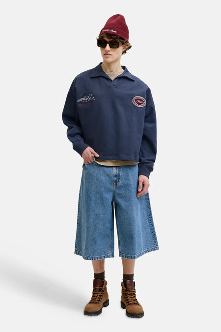 Donkerblauwe sweater met polokraag van ORIGINALS BY JACK & JONES met geborduurde logo's, gestyled met een rode muts, zonnebril, denim shorts en bruine laarzen.