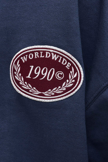 Pull bleu marine à col rond de ORIGINALS BY JACK & JONES, avec un patch brodé bordeaux portant l'inscription 'WORLDWIDE 1990©'.