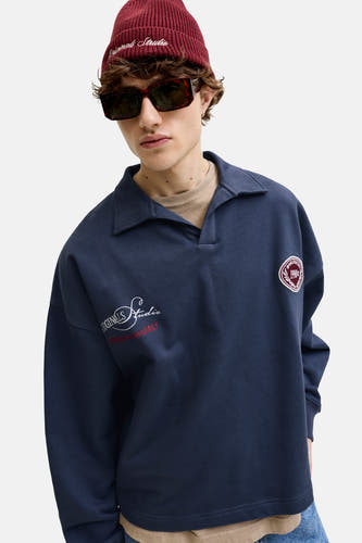Pull bleu marine avec col de ORIGINALS BY JACK & JONES, avec logos brodés blancs et rouges sur la manche et la poitrine, porté par un homme avec un bonnet rouge et des lunettes de soleil.