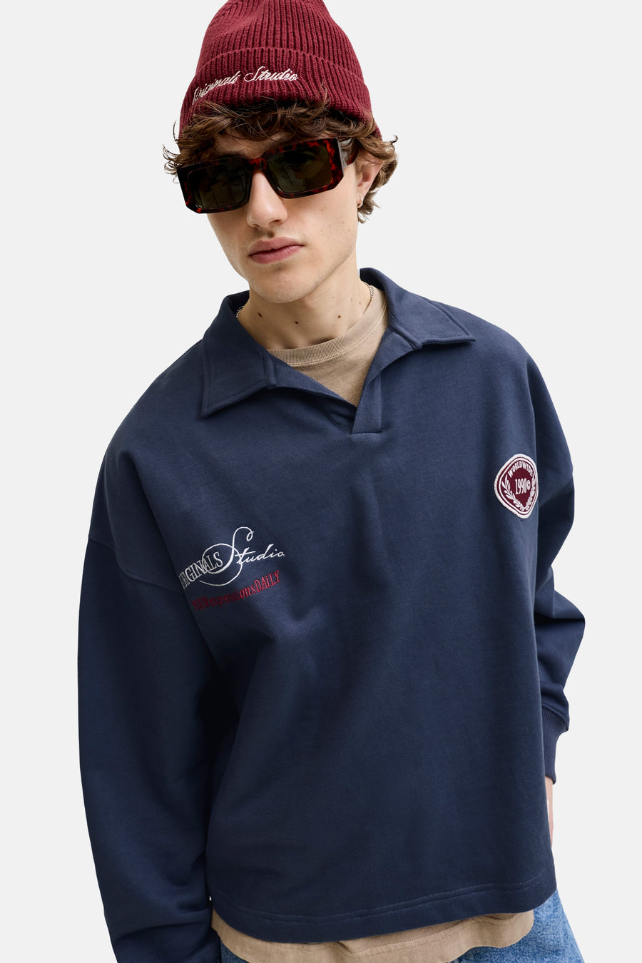 Pull bleu marine avec col de ORIGINALS BY JACK & JONES, avec logos brodés blancs et rouges sur la manche et la poitrine, porté par un homme avec un bonnet rouge et des lunettes de soleil.