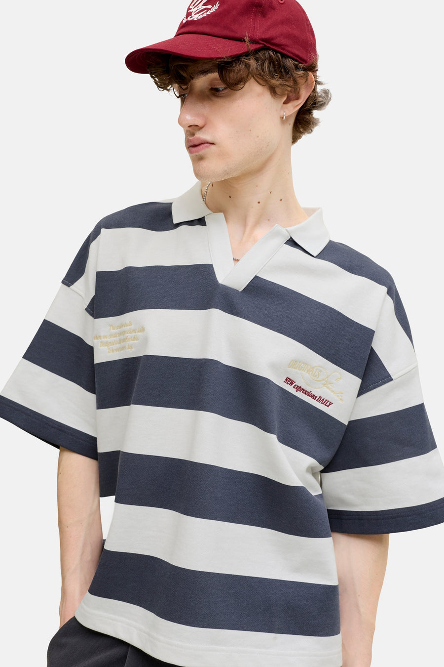 Pull rayé bleu marine et blanc cassé de ORIGINALS BY JACK & JONES, avec col polo, encolure en V et broderie sur la poitrine. Modèle avec casquette rouge.
