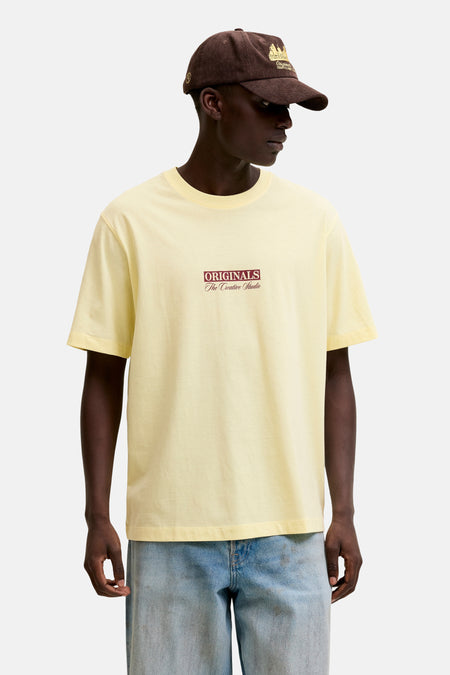 T-shirt à manches courtes jaune de ORIGINALS BY JACK & JONES avec imprimé graphique, porté avec une veste en cuir noire et un jean ample.