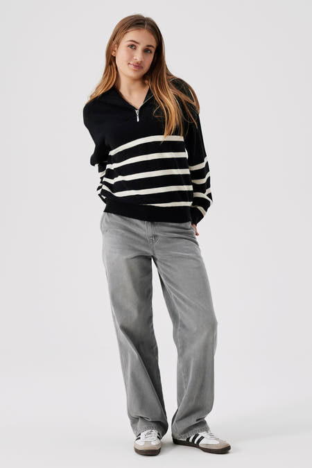 Marinière noir et blanc rayée de LMTD, avec une fermeture éclair et associée à des jeans gris et des baskets.