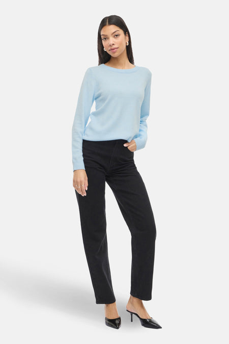 Vrouw met blauwe trui en zwarte broek, zwarte schoenen, witte achtergrond.
