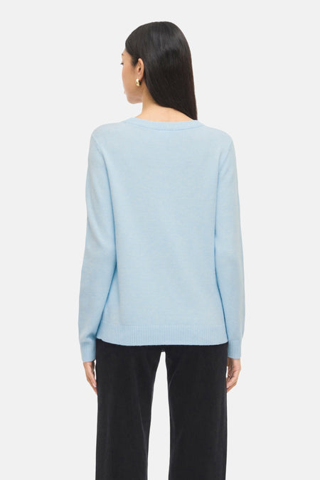 Achterkant van een vrouw in een lichtblauwe trui en zwarte broek.