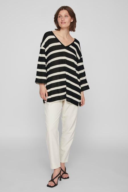 Pull noir à col V de VILA, avec des rayures horizontales et porté avec un pantalon blanc et des sandales noires.
