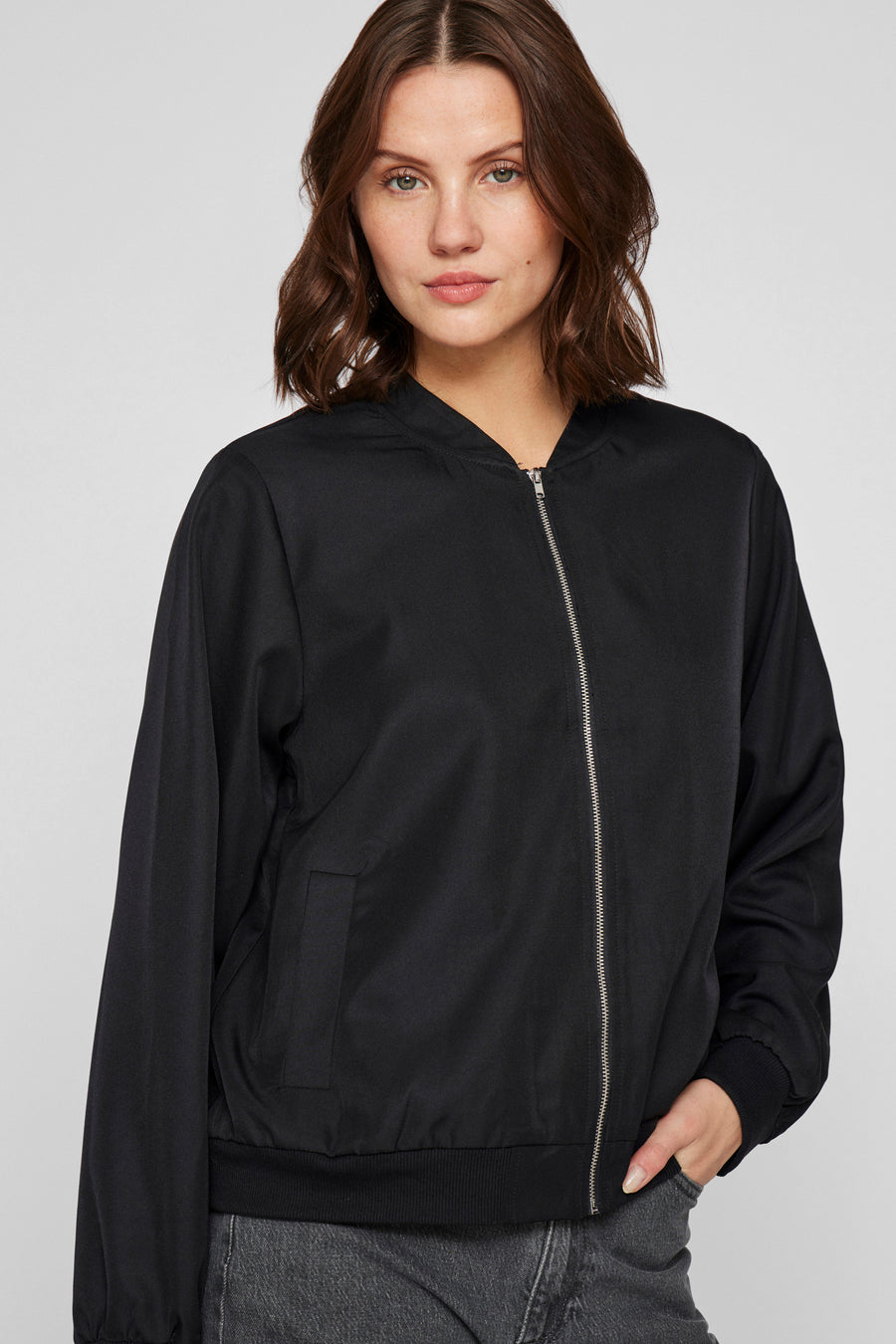 Manteau court noir de VILA avec des manches longues, une fermeture éclair et des poches, porté avec un jean gris.
