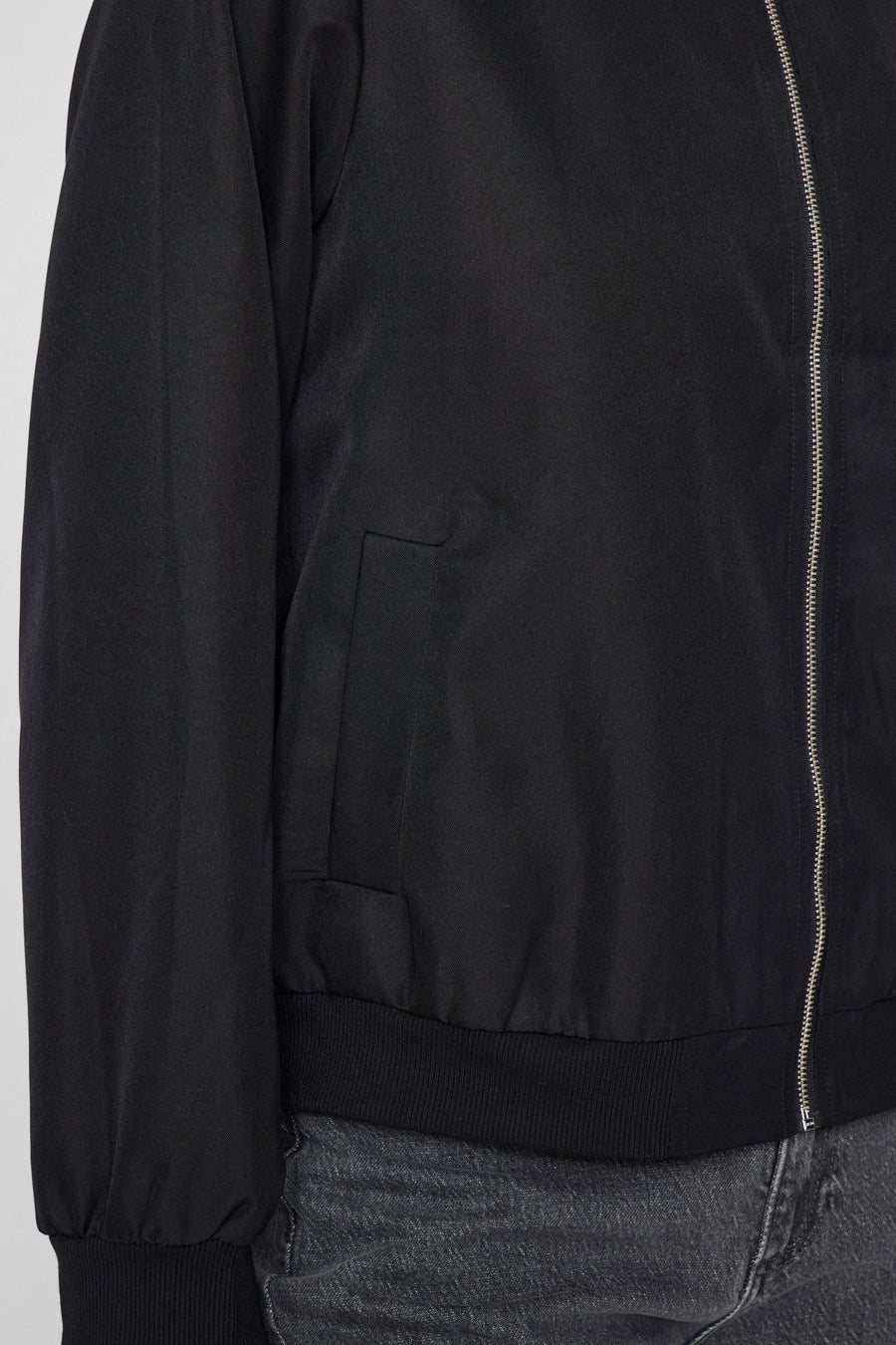 Manteau court noir de VILA, avec fermeture à glissière et poche poitrine, porté sur un jean gris.