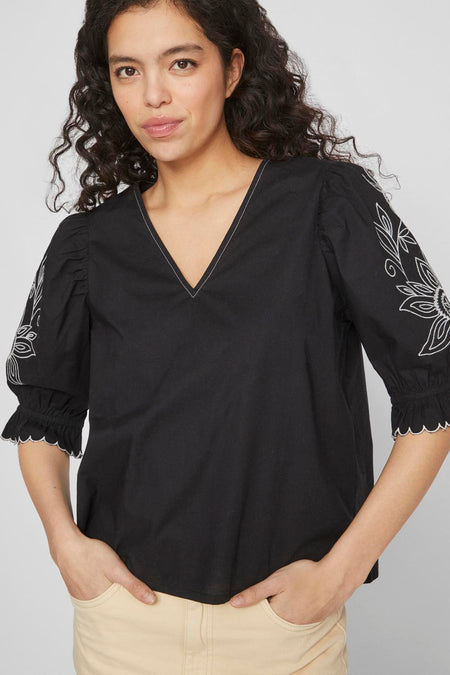 Blouse noire de VILA avec des manches trois-quarts, des fleurs brodées sur les manches et un col en V avec une patte blanche.