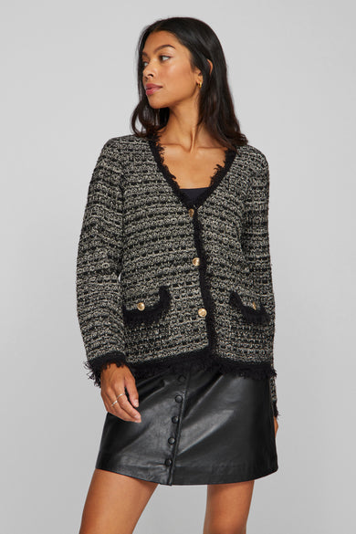 Gilet noir de VILA®, avec fermeture à boutons, poches et bordures froncées, porté avec une jupe en cuir et des chaussettes jusqu'aux genoux.